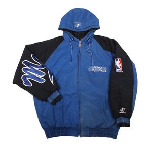Op de afbeelding: Blauwe en zwarte jas met capuchon en een volledige ritssluiting. De jas heeft het Orlando Magic-logo en het NBA-embleem. De mouwen hebben een wit en blauw ontwerp. De jas heeft een capuchon met trekkoord en een logo op de capuchon.