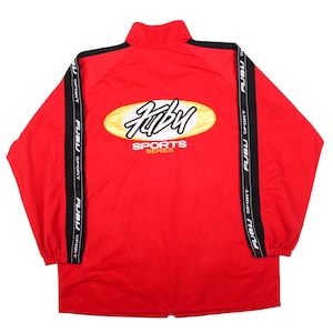 May include: A bright red windbreaker jacket featuring a high collar and long sleeves with black side stripes. The stripes are printed with the white text FUBU SPORT. A large yellow oval logo with the black text FUBU and SPORTS SERIES is on the back.