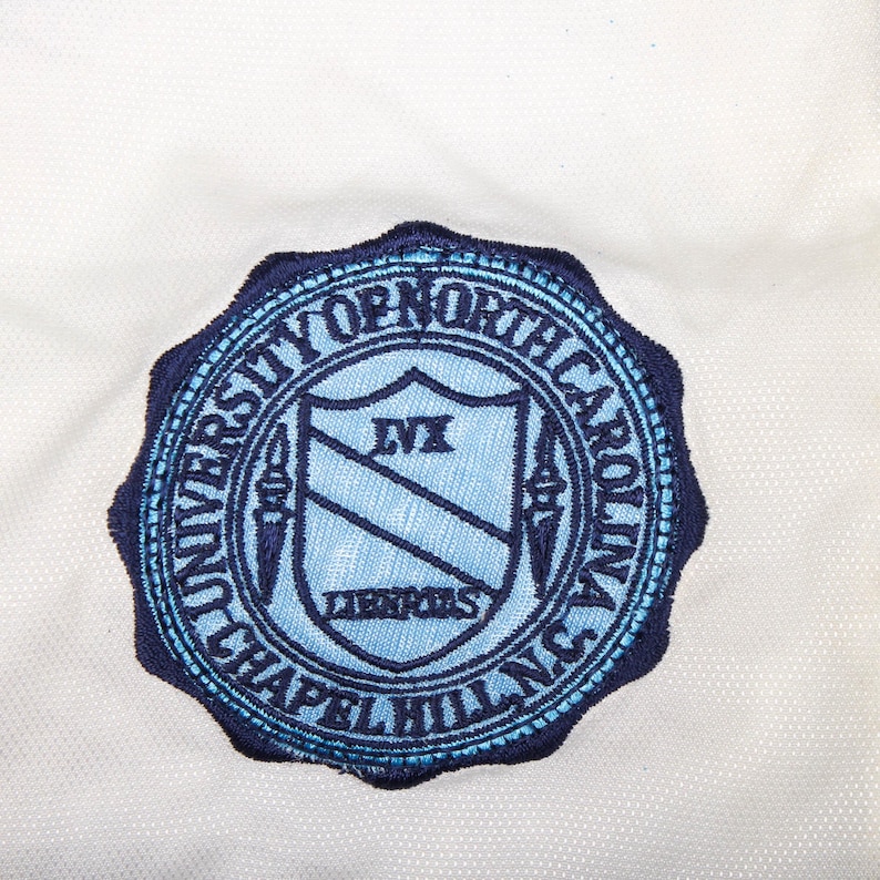 Op de afbeelding: Een marineblauw en lichtblauw geborduurd embleem met de woorden "UNIVERSITY OF NORTH CAROLINA CHAPEL HILL N.C." rondom de rand. Het midden heeft een schildontwerp met de letters "LVI" en het woord "LIBRARIES".