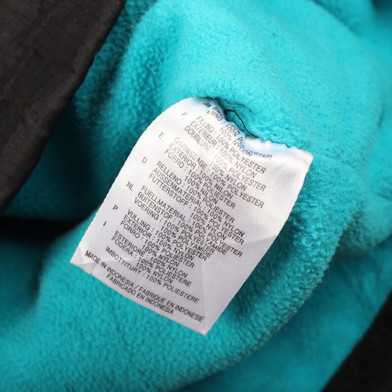 May include: A close-up shot of a clothing label with text in multiple languages, including English, French, and Spanish. The label is white with black text and is attached to a teal blue fabric. The label details the materials.