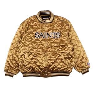 May include: A gold quilted satin jacket featuring the word SAINTS embroidered in black and white across the chest. The jacket has a ribbed collar and cuffs with black, white, and gold stripes, a full-length front zipper, and an NFL logo patch.