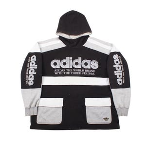 Op de afbeelding: Zwarte, witte en grijze Adidas hoodie. De sweatshirt heeft het Adidas-logo op de borst, de tekst "ADIDAS THE WORLD BRAND WITH THE THREE STRIPES" en het Adidas-logo op de mouwen. Het heeft twee voorzakken.