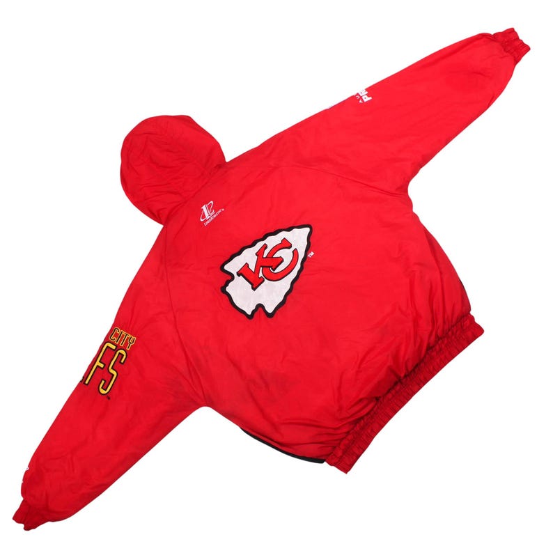May include: A bright red long-sleeve jacket featuring a white arrowhead logo with red KC letters on the back. The sleeve displays the text CITY FS in yellow and red. The jacket includes a hood, elastic cuffs, and a hem, with additional branding logos.