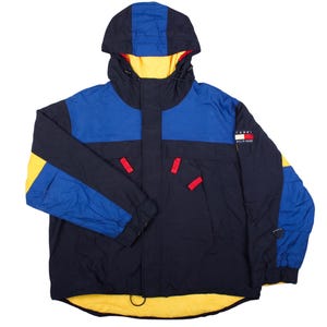 May include: A colour-blocked hooded jacket in navy blue, royal blue, and yellow. The jacket features a front zip closure with a storm flap, red pull tabs on the pockets, and a Tommy Hilfiger logo patch on the left sleeve. The interior lining is yellow.