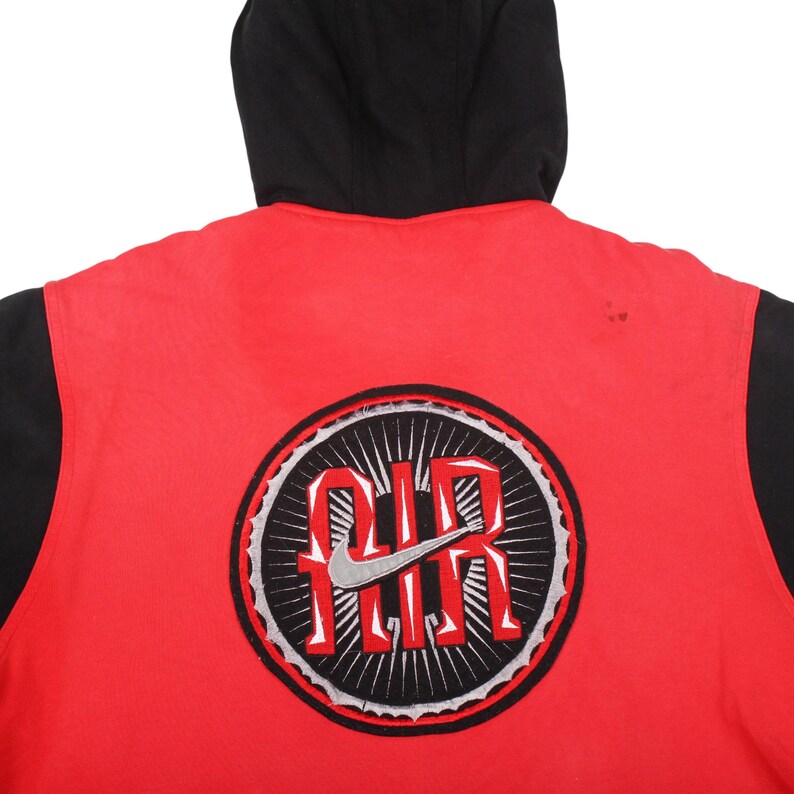 Pu&ograve; includere: Giacca con cappuccio rossa e nera con un grande logo circolare sul retro. Il logo presenta la parola "AIR" in bianco, con un logo Nike argentato, circondato da un design in bianco e nero.
