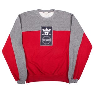 May include: A grey and red crewneck sweatshirt featuring a central rectangular navy blue patch with a white Adidas trefoil logo and the text "DIE WELTMARKE MIT DEN 3 STREIFEN THE BRAND". The garment has long sleeves with grey and red color blocking.