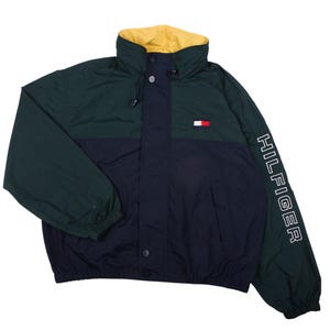 May include: A color-blocked jacket featuring a dark green upper section and a navy blue lower section. The jacket has a yellow inner collar, a front zipper closure with snap buttons, and the word HILFIGER printed in white on the left sleeve.