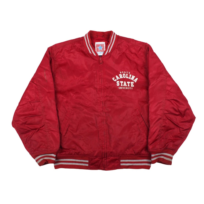 vintage 90s red nylon North Carolina State University varsity bomber jacket on a white background, featuring red and grey striped athletic rib-knit collar, cuffs, and hem, with an arched white text print of the school name on the left chest.