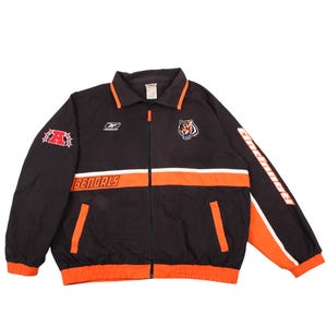 May include: A black Reebok zip-up jacket featuring orange and white horizontal stripes, a tiger head logo, and the text BENGALS and Cincinnati. The right sleeve displays a red letter A surrounded by stars, with orange cuffs and a collar trim.