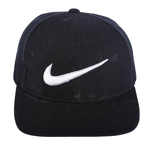 Puede incluir: Una gorra de béisbol negra con un prominente logotipo Nike swoosh bordado en blanco en el panel frontal. La gorra tiene una corona estructurada, una visera plana y ojales cosidos para la ventilación, con un diseño deportivo clásico.