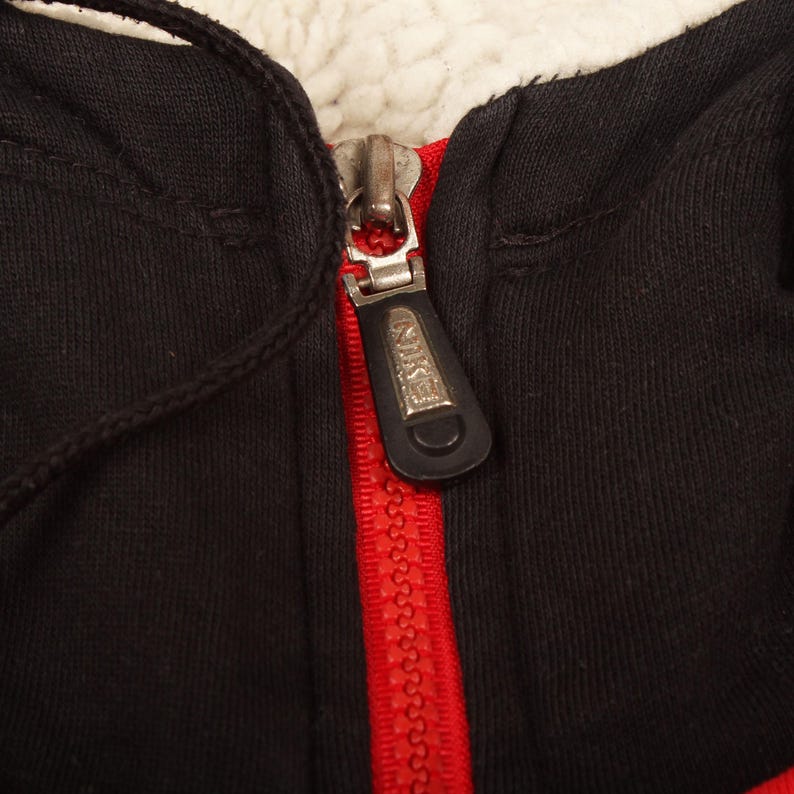 Pu&ograve; includere: Primo piano di una felpa con cappuccio nera con cerniera rossa e fodera in pile bianco. Il tirazip ha il logo Nike. Il cordino &egrave; nero.