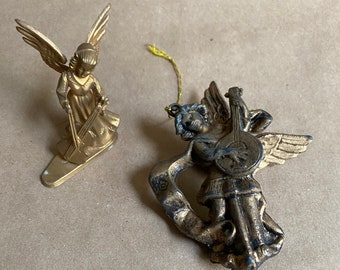 May include: Two vintage gold-coloured angel ornaments. One angel is playing a violin, and the other is playing a stringed instrument. Both angels have wings and are made of metal. The ornaments are on a neutral-coloured surface.