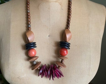 May include: A wooden beaded necklace with large, colorful beads. The necklace features red, black, and brown wooden beads, as well as a cluster of pink wooden beads.