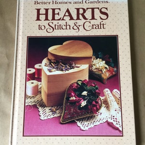 May include: A book titled "Hearts to Stitch & Craft" by Better Homes and Gardens. The cover features a heart-shaped wooden box with a heart-shaped fabric pillow inside. The box is filled with dried flowers and there are spools of thread and a crocheted doily on the table.