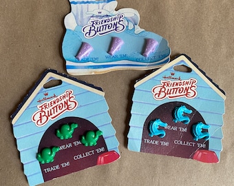 May include: Three sets of Hallmark Friendship Buttons. Each set includes three plastic buttons with different designs. The first set features purple ice cream cones, the second set features green dinosaurs, and the third set features blue alligators. Each card has the text "Hallmark Friendship Buttons" and "Trade 'em! Wear 'em! Collect 'em!"