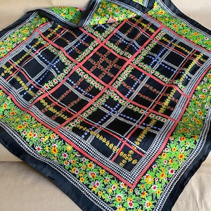 Puede incluir: Bufanda de seda negra con un patrón a cuadros y un borde floral. La bufanda tiene un patrón a cuadros rojo, blanco y azul con un fondo negro. El borde es un patrón floral con hojas verdes y flores amarillas, rojas y blancas.