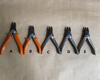 May include: Five different wire cutters are displayed, labeled A through E. Cutters A and B have orange handles, while C, D, and E have silver handles. All have black cutting blades and are arranged on a brown surface.