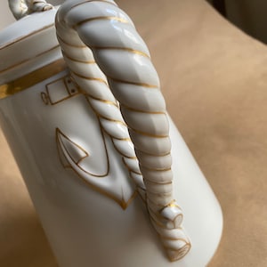 May include: White ceramic nautical-themed pitcher with gold accents. The handle is designed to resemble a rope. Features an anchor and other nautical details in gold. The lid has a small decorative element.