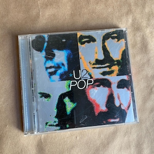 May include: A CD case with a colorful image of the band U2 on the front. The band members are depicted in a pop art style with their faces in close-up. The title of the album, "U2 POP", is printed in black and white letters on the front of the case.