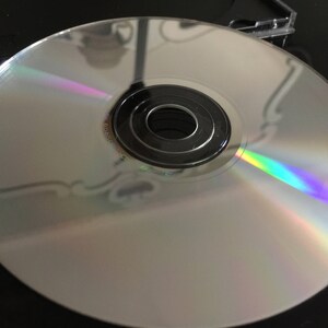 Puede incluir: Un disco compacto plateado con un brillo iridiscente arco&iacute;ris. El disco tiene un agujero central y un anillo negro alrededor.
