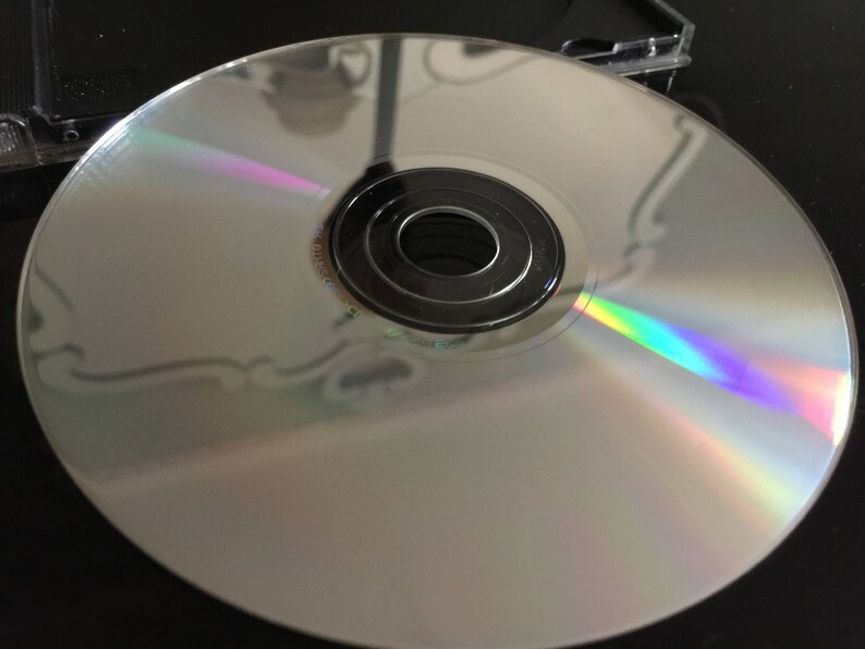 Puede incluir: Un disco compacto plateado con un brillo iridiscente arco&iacute;ris. El disco tiene un agujero central y un anillo negro alrededor.