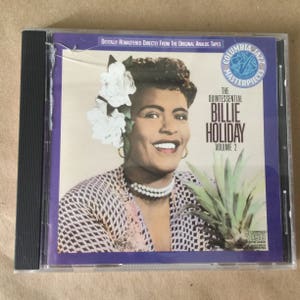May include: A CD case with a purple background featuring a photo of Billie Holiday, a famous jazz singer. The text on the case reads "The Quintessential Billie Holiday, Volume 2".