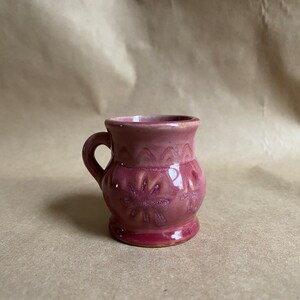 May include: A small, pink ceramic mug with a handle and a floral design. The mug is sitting on a brown surface.