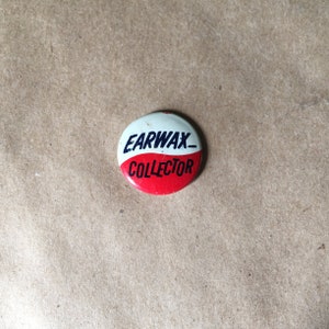 May include: A red and white button with the text "Earwax Collector" in black lettering.