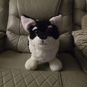 May include: A plush dog toy with black and white fur, pink inner ears, and a black nose. The stuffed animal is sitting on a gray leather couch.