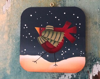 May include: A hand-painted wooden ornament featuring a red cardinal wearing a green and white striped scarf, standing on a snowy landscape against a dark blue background with white snowflakes.