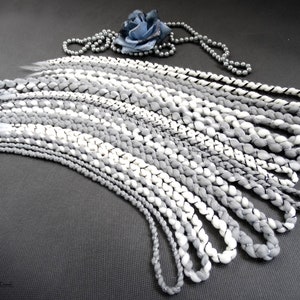 Puede incluir: Un conjunto de extensiones de cabello trenzadas grises y blancas con un hilo negro que recorre cada trenza. Las extensiones están dispuestas en forma de abanico sobre una superficie gris.