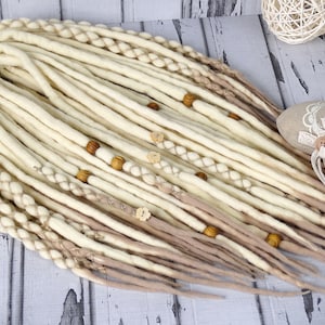 Puede incluir: Un conjunto de rastas largas de lana blanca y beige con cuentas de madera. Las rastas están dispuestas en círculo sobre una superficie de madera blanca.