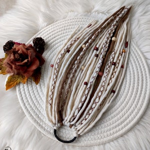 May include: A set of handmade dreadlocks in shades of white, brown, and beige, adorned with wooden and red beads. The dreadlocks are secured with a black elastic band. The image is set on a white woven mat and a fluffy white surface.