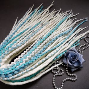 Puede incluir: Dreadlocks sintéticas blancas y azules con cuentas plateadas. Las dreadlocks están dispuestas en círculo y se acentúan con un collar de perlas plateadas y blancas y una rosa de tela azul.