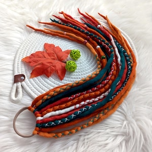 Puede incluir: Una colección de rastas naranjas, rojas y verde azulado unidas a una goma para el pelo marrón. Las rastas están decoradas con costuras blancas y verde azulado. Una hoja decorativa naranja y dos bolas de hilo verde se encuentran sobre un posavasos blanco tejido.