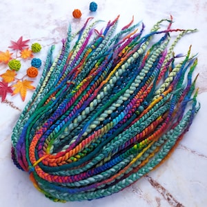 May include: A bundle of vibrant, braided dreadlocks in a spectrum of colours including blue, green, purple, and orange. The dreadlocks display varied textures and colour gradients. Decorative balls and autumn leaves are scattered around the dreadlocks.