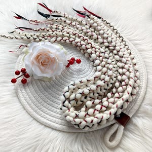 Può includere: Un accessorio per capelli intrecciati bianchi con dettagli rossi e neri, una rosa bianca e bacche rosse. L'accessorio è posto su un tappetino rotondo bianco intrecciato con manico in pelle e corda.