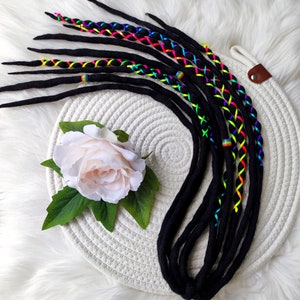 May include: Black dreadlocks with vibrant rainbow-colored accents. The dreads are adorned with colorful thread woven in a crisscross pattern. A white flower with green leaves rests near the dreadlocks, which are displayed on a white woven mat.