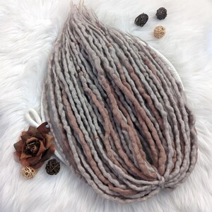 May include: A bundle of handmade dreadlocks in shades of gray and brown, with a gradient effect. The dreadlocks are arranged on a white, fluffy surface. Decorative elements include a brown fabric flower and small woven spheres.