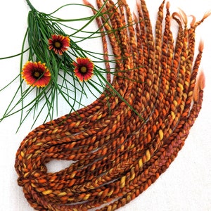 Puede incluir: Un conjunto de extensiones de cabello sintético con textura de trenza retorcida en una paleta de colores variados de naranja intenso, amarillo dorado y marrón, acompañado de una rama decorativa de hierba verde con tres pequeñas flores rojas y amarillas.