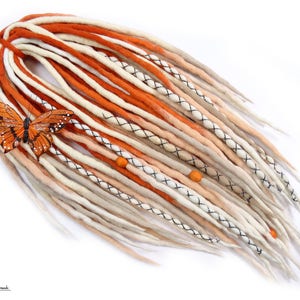 Puede incluir: Un conjunto de rastas sintéticas de color naranja, melocotón y blanco con detalles negros y cuentas naranjas. Las rastas están dispuestas en forma de abanico y un amuleto de mariposa está unido a la parte superior.