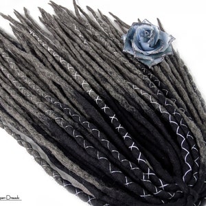 Puede incluir: Un conjunto de rastas grises y negras con detalles de costura blanca en forma de cruz. Una flor azul claro con purpurina adorna las rastas. El logotipo "Super Dreads" es visible en la esquina inferior izquierda.