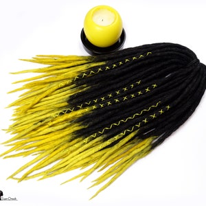 Puede incluir: Dreadlocks sintéticas ombre negro y amarillo con costuras amarillas. Las dreadlocks están dispuestas en forma de abanico sobre un fondo blanco.