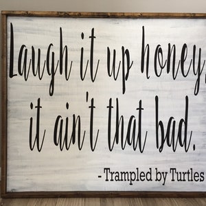May include: White wooden sign with black text that reads "Laugh it up honey, it ain't that bad. - Trampled by Turtles".