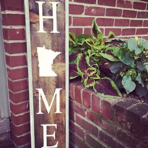 Puede incluir: Un letrero de madera con un marco blanco y la palabra "HOME" escrita en letras blancas. La letra "O" está reemplazada por el contorno del estado de Minnesota.