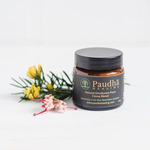 May include: A small glass jar with a black lid containing natural deodorant paste. The jar has a black label with the text "Paudha Healing" and "Natural Deodorant Paste Citrus Blend" in white. The jar is sitting on a white surface with a sprig of yellow and pink flowers.