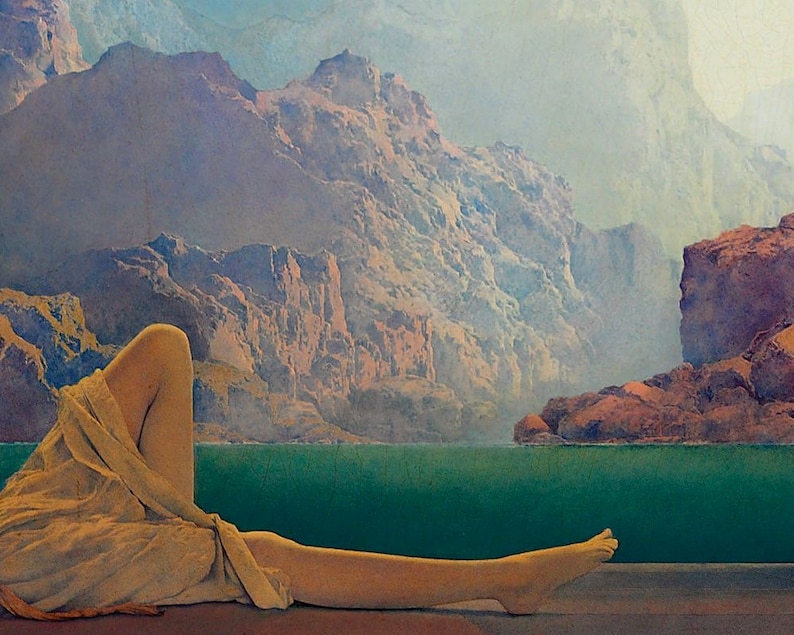May include: A woman's leg and foot are visible in the foreground, draped in a flowing beige fabric. The background features a scenic landscape with a turquoise body of water and a hazy mountain range in the distance.