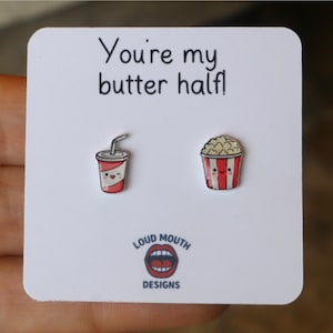 May include: A pair of novelty earrings on a white card. One earring is a red and white soda cup with a straw, and the other is a red and white popcorn container. The card reads "You're my butter half!" and features the "Loud Mouth Designs" logo.