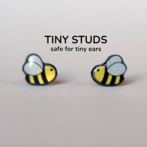 May include: A pair of tiny bee stud earrings. Each earring features a yellow body with black stripes, a light blue wing, and a dark blue outline. The text "TINY STUDS safe for tiny ears" is above the earrings.