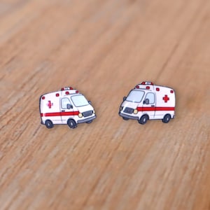 May include: Two white and red ambulance earrings with black wheels and a red cross on the side. The earrings are set against a light brown wooden surface.